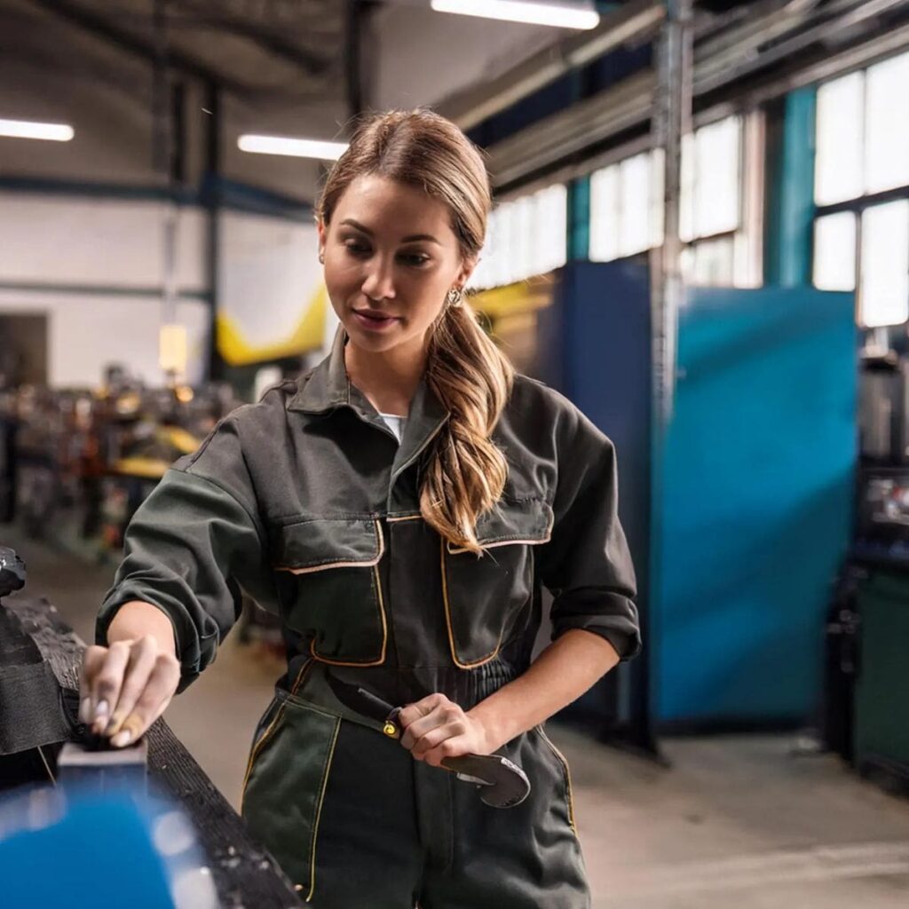 Mujer con mono de trabajo utilizando herramienta en taller industrial, representando ropa laboral moderna y funcional.